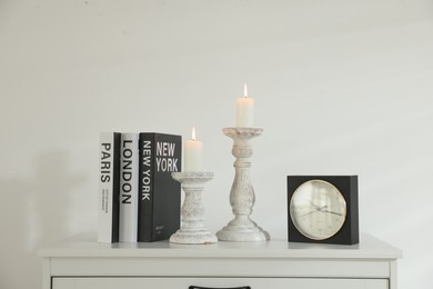 Photo of Stylish candlesticks with burning candles, alarm clock and books on cabinet near white wall indoors