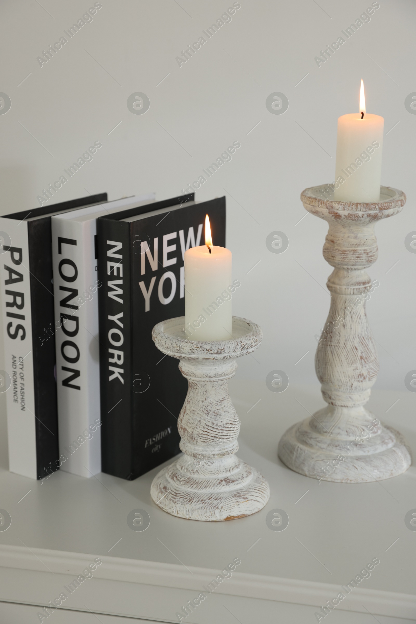 Stylish candlesticks with burning candles, alarm clock and books on cabinet near white wall indoors Photo of Stylish candlesticks with burning candles, alarm clock and books on cabinet near white wall indoors