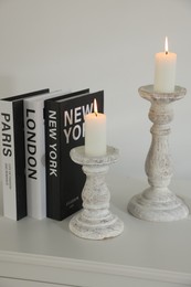 Photo of Stylish candlesticks with burning candles, alarm clock and books on cabinet near white wall indoors