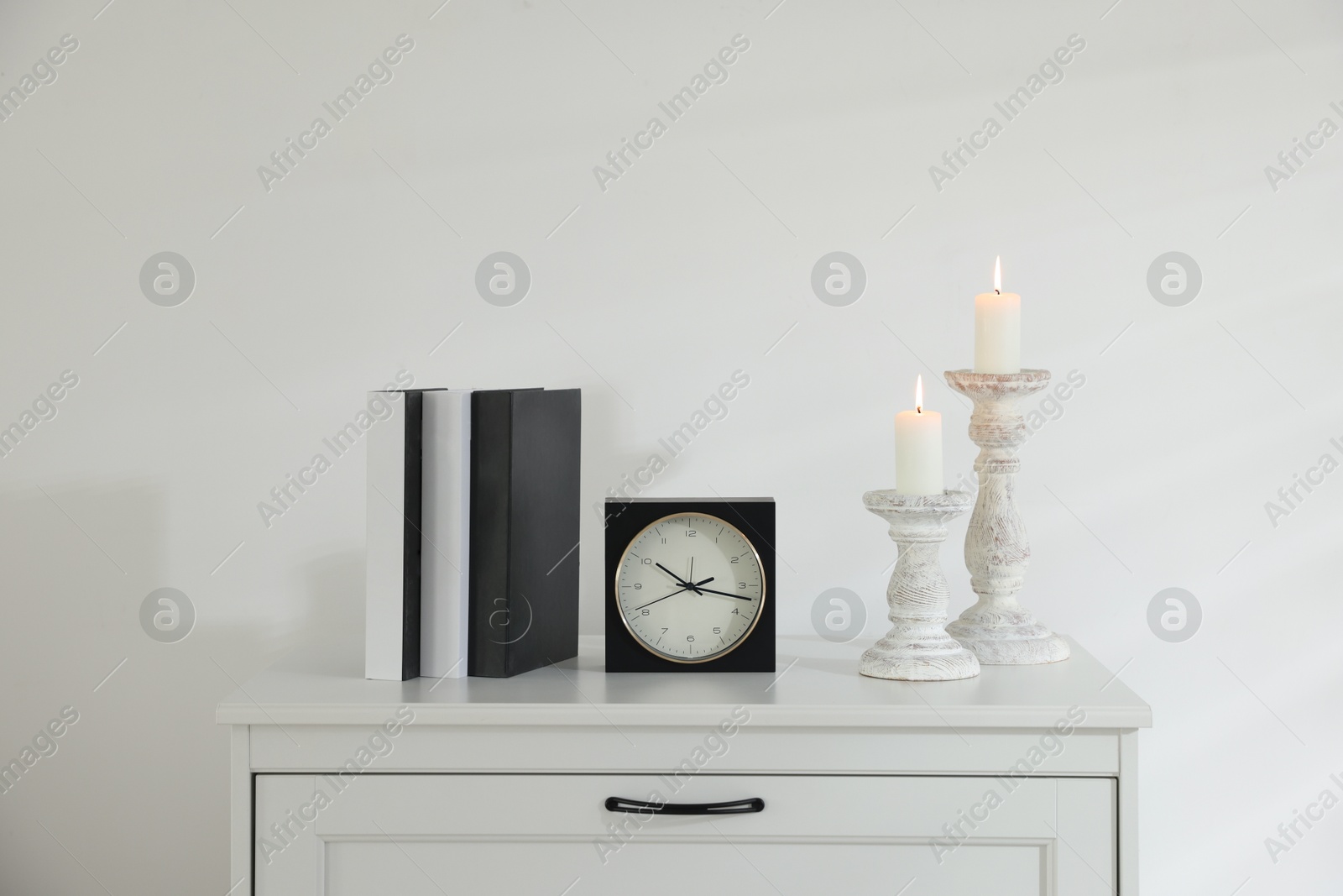 Stylish candlesticks with burning candles, alarm clock and books on cabinet near white wall indoors Photo of Stylish candlesticks with burning candles, alarm clock and books on cabinet near white wall indoors