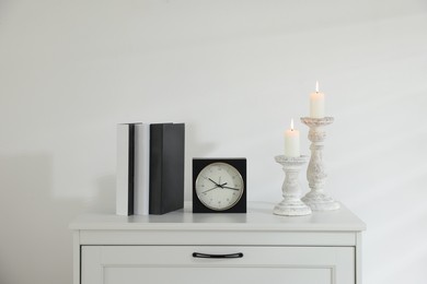 Photo of Stylish candlesticks with burning candles, alarm clock and books on cabinet near white wall indoors