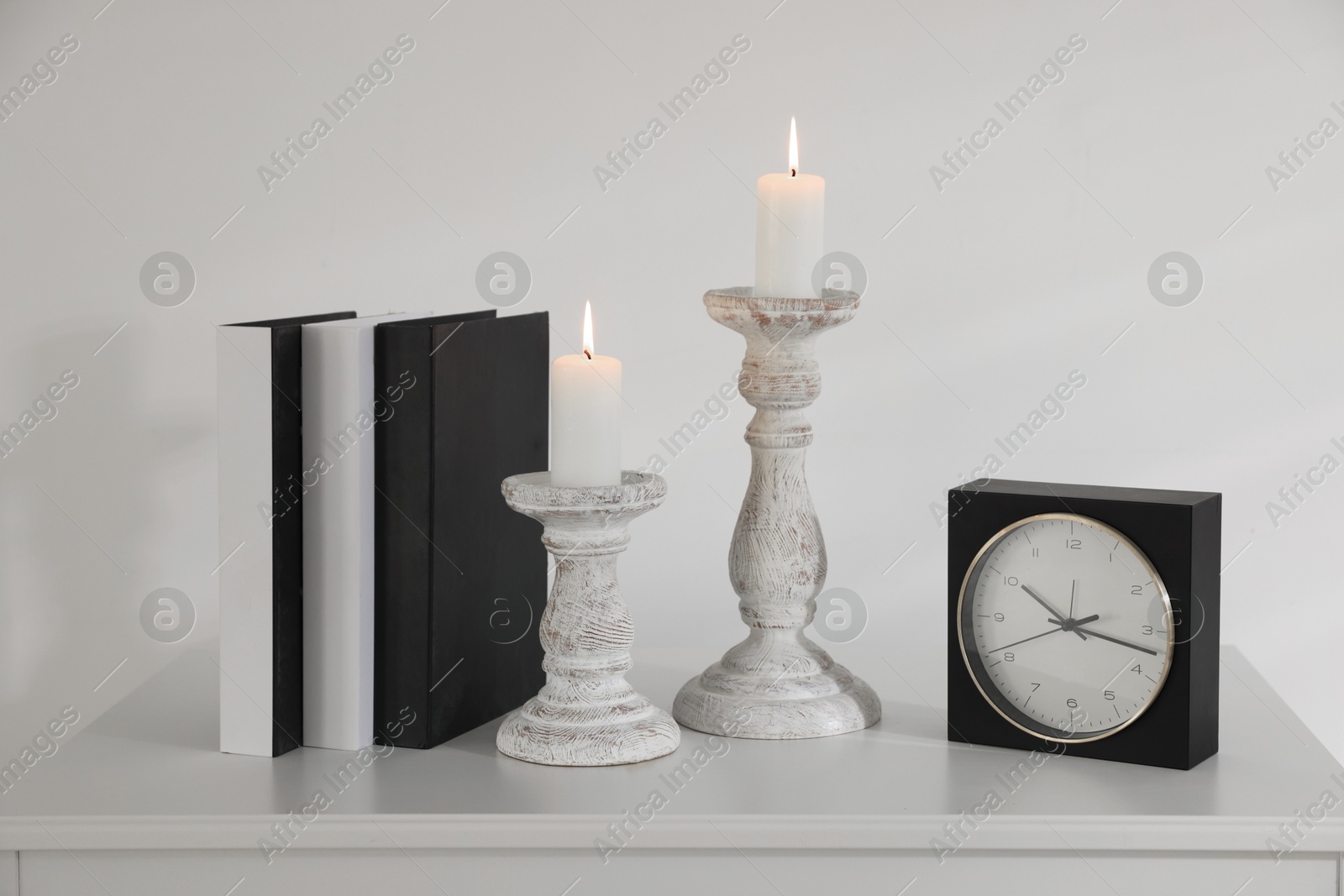 Stylish candlesticks with burning candles, alarm clock and books on cabinet near white wall indoors Photo of Stylish candlesticks with burning candles, alarm clock and books on cabinet near white wall indoors