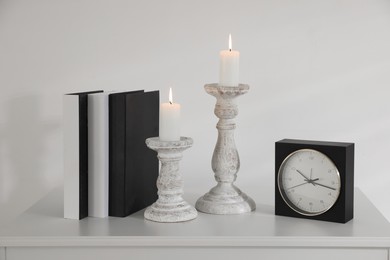 Photo of Stylish candlesticks with burning candles, alarm clock and books on cabinet near white wall indoors