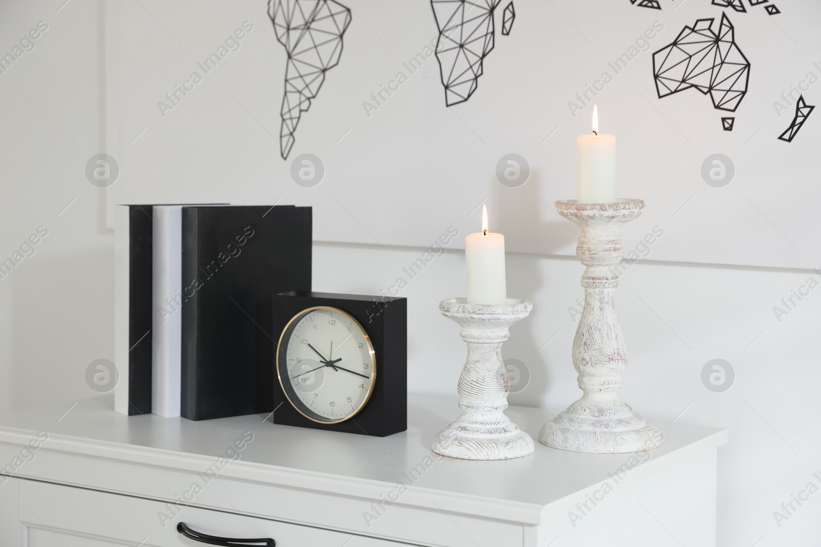 Stylish candlesticks with burning candles, alarm clock and books on cabinet near white wall indoors Photo of Stylish candlesticks with burning candles, alarm clock and books on cabinet near white wall indoors