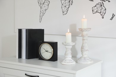 Photo of Stylish candlesticks with burning candles, alarm clock and books on cabinet near white wall indoors