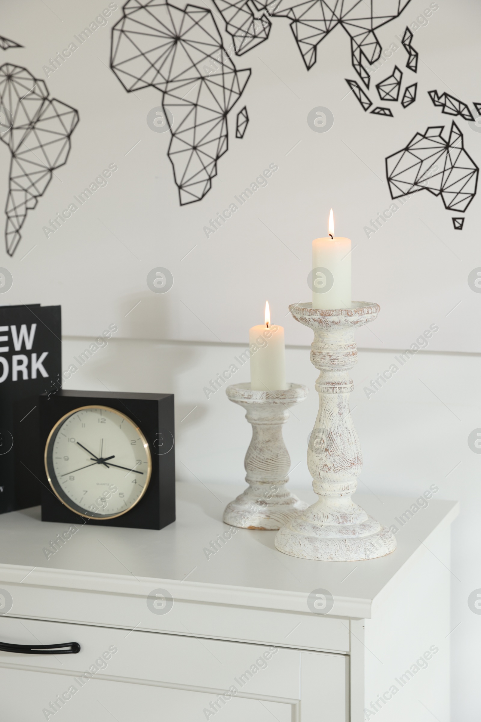 Photo of Stylish candlesticks with burning candles, alarm clock and book on cabinet near white wall indoors