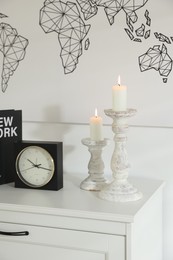 Photo of Stylish candlesticks with burning candles, alarm clock and book on cabinet near white wall indoors