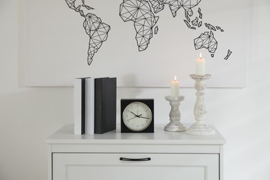 Photo of Stylish candlesticks with burning candles, alarm clock and books on cabinet near white wall indoors