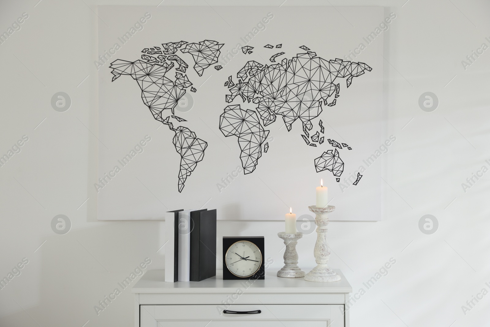 Stylish candlesticks with burning candles, alarm clock and books on cabinet near white wall indoors Photo of Stylish candlesticks with burning candles, alarm clock and books on cabinet near white wall indoors