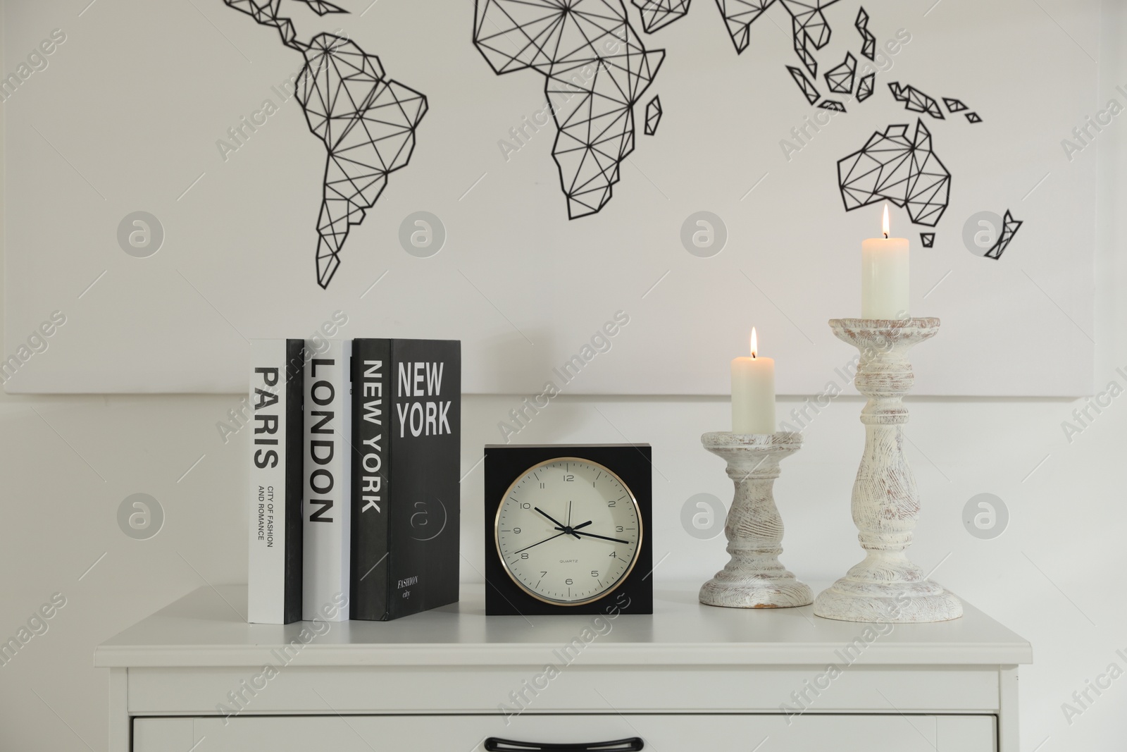 Stylish candlesticks with burning candles, alarm clock and books on cabinet near white wall indoors Photo of Stylish candlesticks with burning candles, alarm clock and books on cabinet near white wall indoors