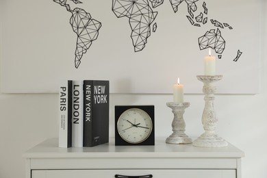 Photo of Stylish candlesticks with burning candles, alarm clock and books on cabinet near white wall indoors