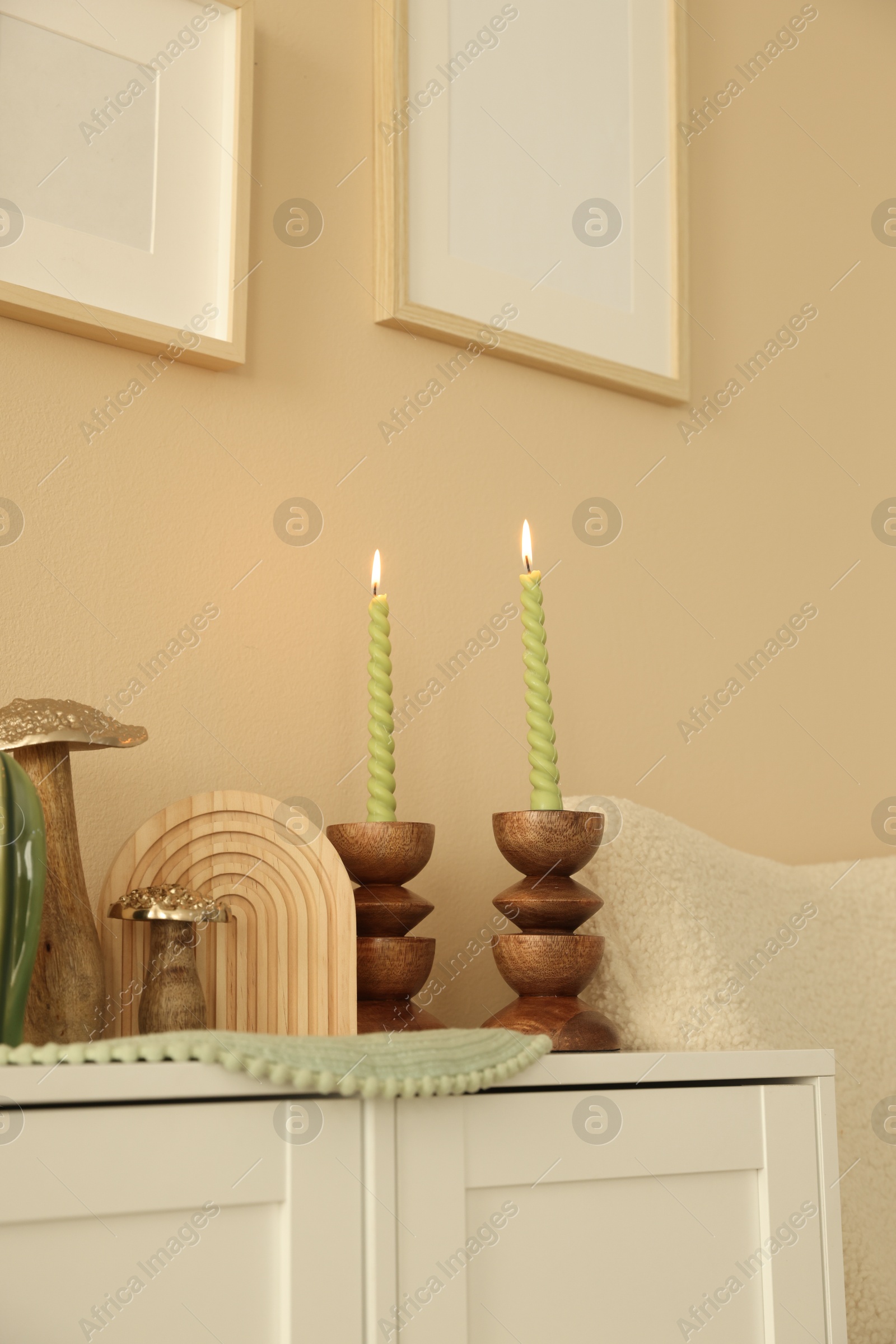 Stylish candlesticks with burning candles and other decor on cabinet near beige wall indoors Photo of Stylish candlesticks with burning candles and other decor on cabinet near beige wall indoors