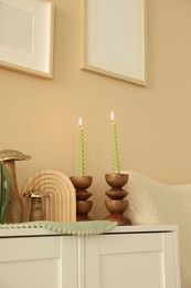 Photo of Stylish candlesticks with burning candles and other decor on cabinet near beige wall indoors
