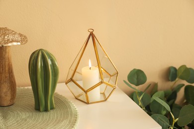 Photo of Burning candle in holder and other decor on cabinet near beige wall indoors