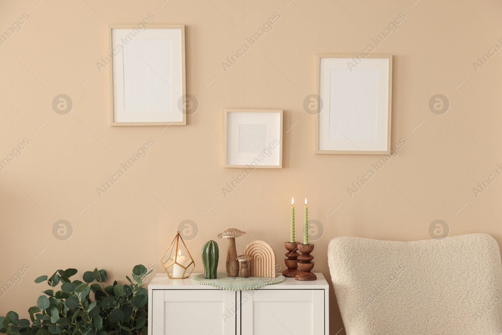Stylish candlesticks with burning candles and other decor on cabinet near beige wall indoors Photo of Stylish candlesticks with burning candles and other decor on cabinet near beige wall indoors