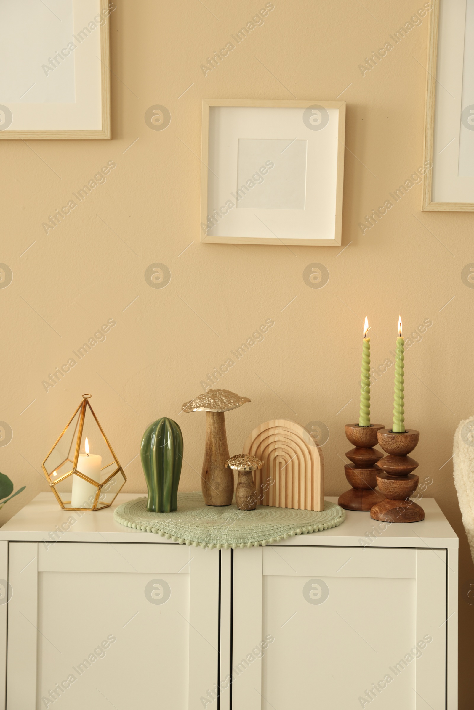Stylish candlesticks with burning candles and other decor on cabinet near beige wall indoors Photo of Stylish candlesticks with burning candles and other decor on cabinet near beige wall indoors