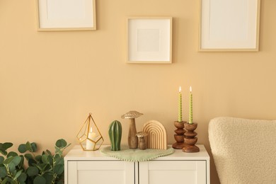 Photo of Stylish candlesticks with burning candles and other decor on cabinet near beige wall indoors