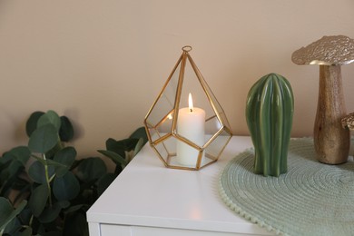 Photo of Burning candle in holder and other decor on cabinet near beige wall indoors