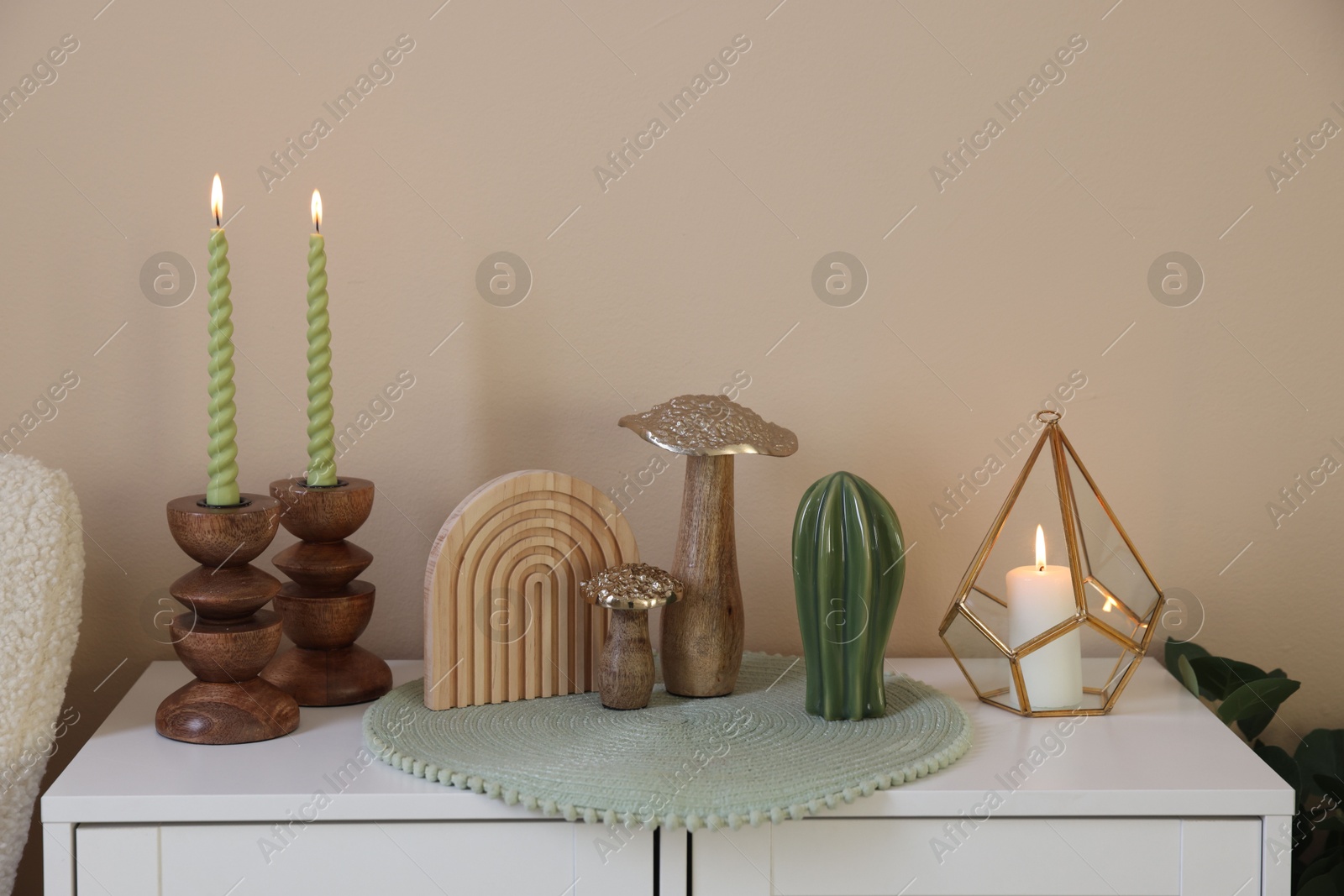 Stylish candlesticks with burning candles and other decor on cabinet near beige wall indoors Photo of Stylish candlesticks with burning candles and other decor on cabinet near beige wall indoors