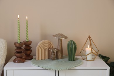 Photo of Stylish candlesticks with burning candles and other decor on cabinet near beige wall indoors