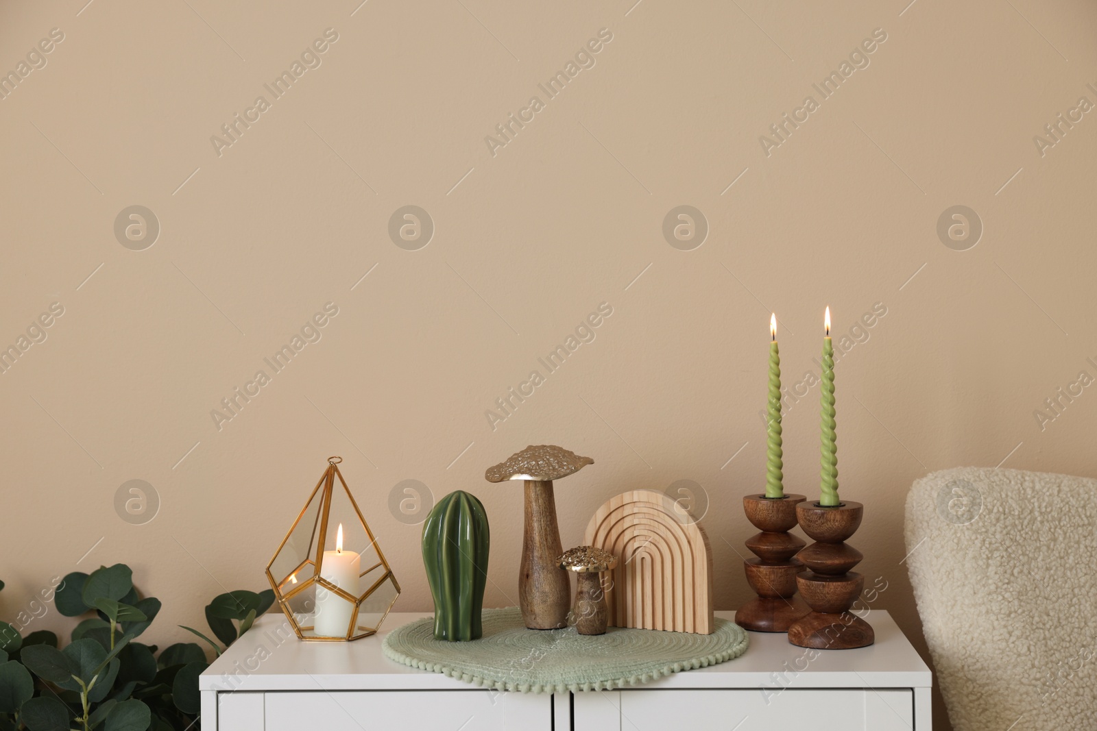 Stylish candlesticks with burning candles and other decor on cabinet near beige wall indoors Photo of Stylish candlesticks with burning candles and other decor on cabinet near beige wall indoors