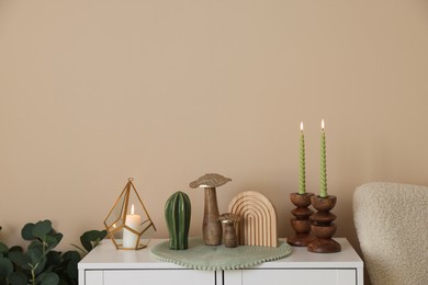 Photo of Stylish candlesticks with burning candles and other decor on cabinet near beige wall indoors