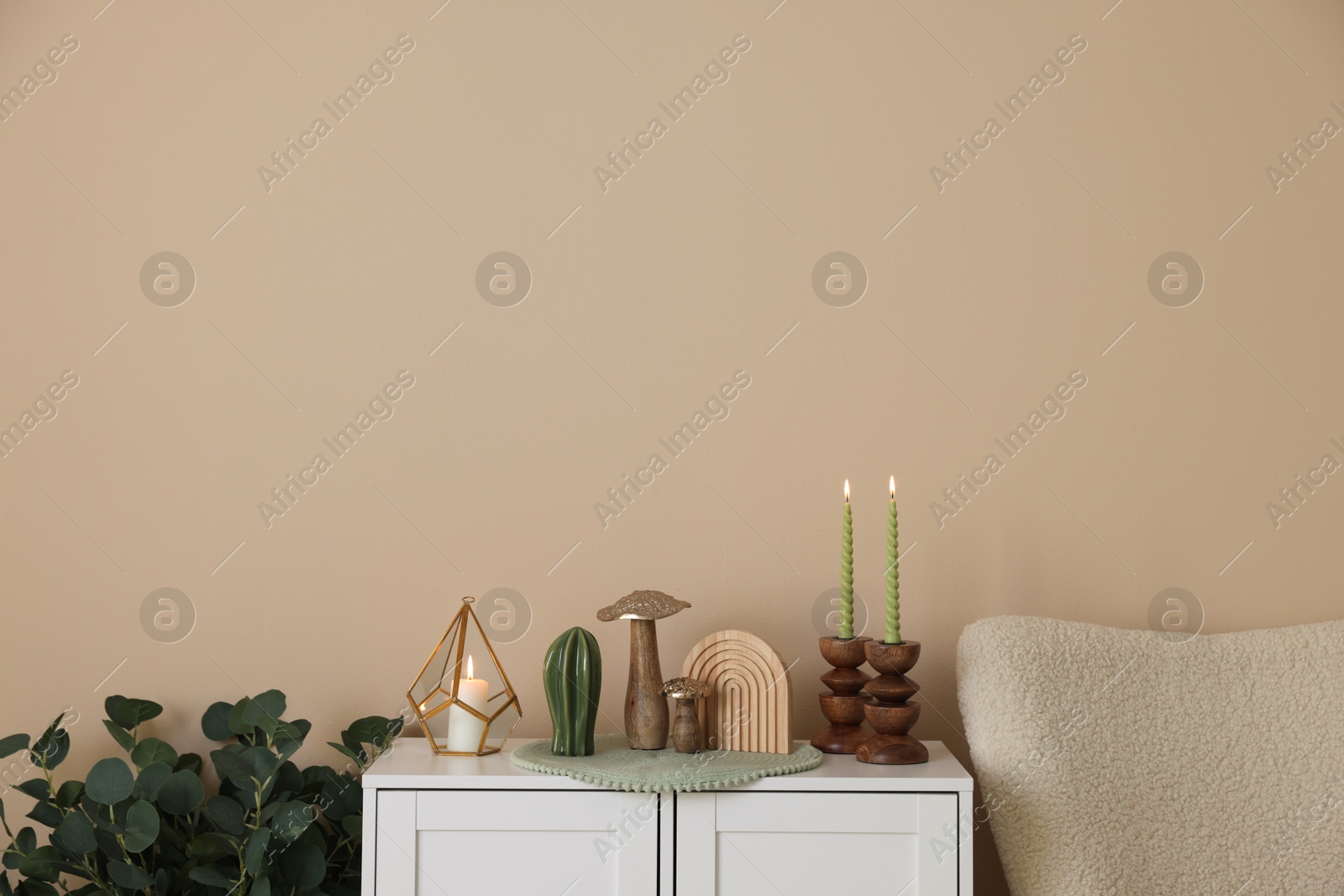 Stylish candlesticks with burning candles and other decor on cabinet near beige wall indoors Photo of Stylish candlesticks with burning candles and other decor on cabinet near beige wall indoors