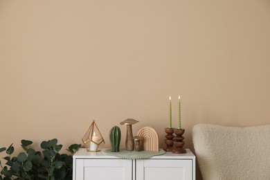 Photo of Stylish candlesticks with burning candles and other decor on cabinet near beige wall indoors