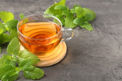 Photo of Aromatic lemon balm tea in glass cup and fresh leaves on gray textured table, closeup. Space for text