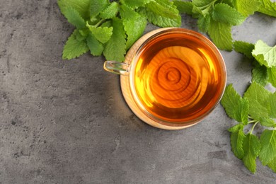 Photo of Aromatic lemon balm tea in glass cup and fresh leaves on gray textured table, flat lay. Space for text