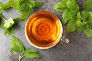 Photo of Aromatic lemon balm tea in glass cup and fresh leaves on gray textured table, flat lay