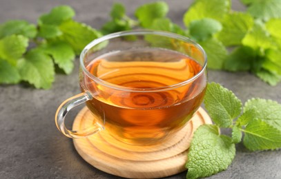 Photo of Aromatic lemon balm tea in glass cup and fresh leaves on gray textured table, closeup