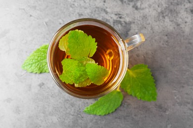 Photo of Aromatic lemon balm tea in glass cup and fresh leaves on gray textured table, top view