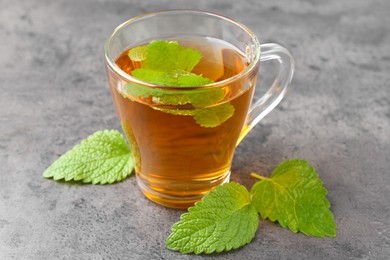 Photo of Aromatic lemon balm tea in glass cup and fresh leaves on gray textured table, closeup