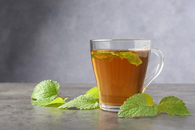 Photo of Aromatic lemon balm tea in glass cup and fresh leaves on gray textured table, space for text