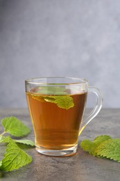 Photo of Aromatic lemon balm tea in glass cup and fresh leaves on gray textured table