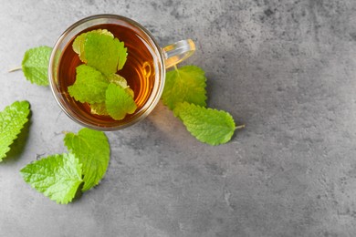Photo of Aromatic lemon balm tea in glass cup and fresh leaves on gray textured table, flat lay. Space for text