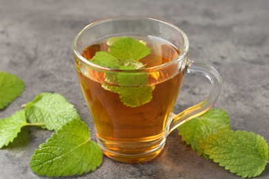 Photo of Aromatic lemon balm tea in glass cup and fresh leaves on gray textured table, closeup