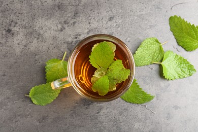 Photo of Aromatic lemon balm tea in glass cup and fresh leaves on gray textured table, flat lay
