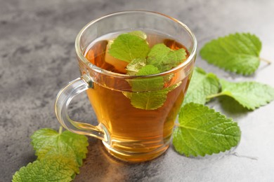 Photo of Aromatic lemon balm tea in glass cup and fresh leaves on gray textured table, closeup