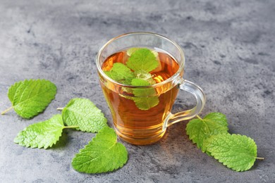 Photo of Aromatic lemon balm tea in glass cup and fresh leaves on gray textured table, closeup