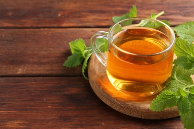 Photo of Aromatic lemon balm tea in glass cup and fresh leaves on wooden table, closeup. Space for text