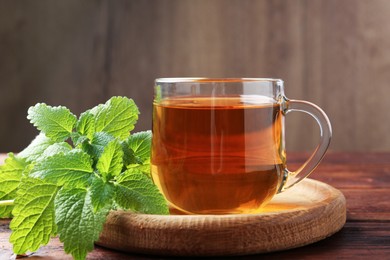 Photo of Aromatic lemon balm tea in glass cup and fresh leaves on wooden table, closeup
