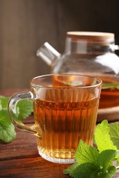 Photo of Aromatic lemon balm tea in glass cup, teapot and fresh leaves on wooden table, closeup