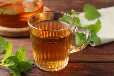 Photo of Aromatic lemon balm tea in glass cup, teapot and fresh leaves on wooden table, closeup