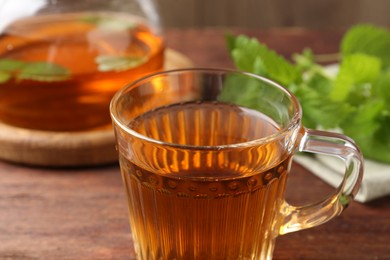 Photo of Aromatic lemon balm tea in glass cup, teapot and fresh leaves on wooden table, closeup