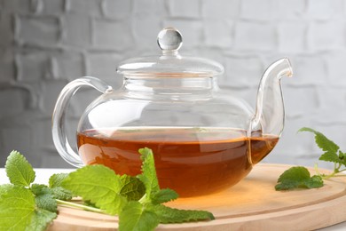 Photo of Aromatic lemon balm tea in glass teapot and fresh leaves on table against light background, closeup
