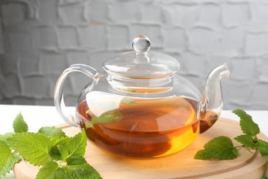 Photo of Aromatic lemon balm tea in glass teapot and fresh leaves on table against light background, closeup