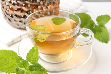 Photo of Aromatic lemon balm tea in glass cup, teapot and fresh leaves on white table, closeup