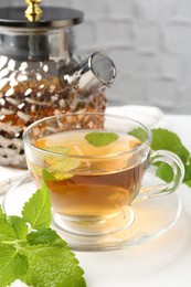 Photo of Aromatic lemon balm tea in glass cup, teapot and fresh leaves on white table, closeup
